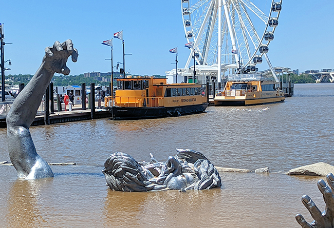 Statue of two giant hands partially submerged in water, symbolizing struggle against rising sea levels, with a ferris wheel and boats in the background at a coastal city waterfront.